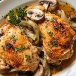 A photo of a dish of Garlic Butter Chicken. The chicken is golden brown and is covered in a garlic butter sauce. There are also some vegetables, such as mushrooms and onions, in the dish. The dish is served on a white plate and is garnished with parsley. The background is a beige surface.