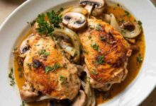 A photo of a dish of Garlic Butter Chicken. The chicken is golden brown and is covered in a garlic butter sauce. There are also some vegetables, such as mushrooms and onions, in the dish. The dish is served on a white plate and is garnished with parsley. The background is a beige surface.